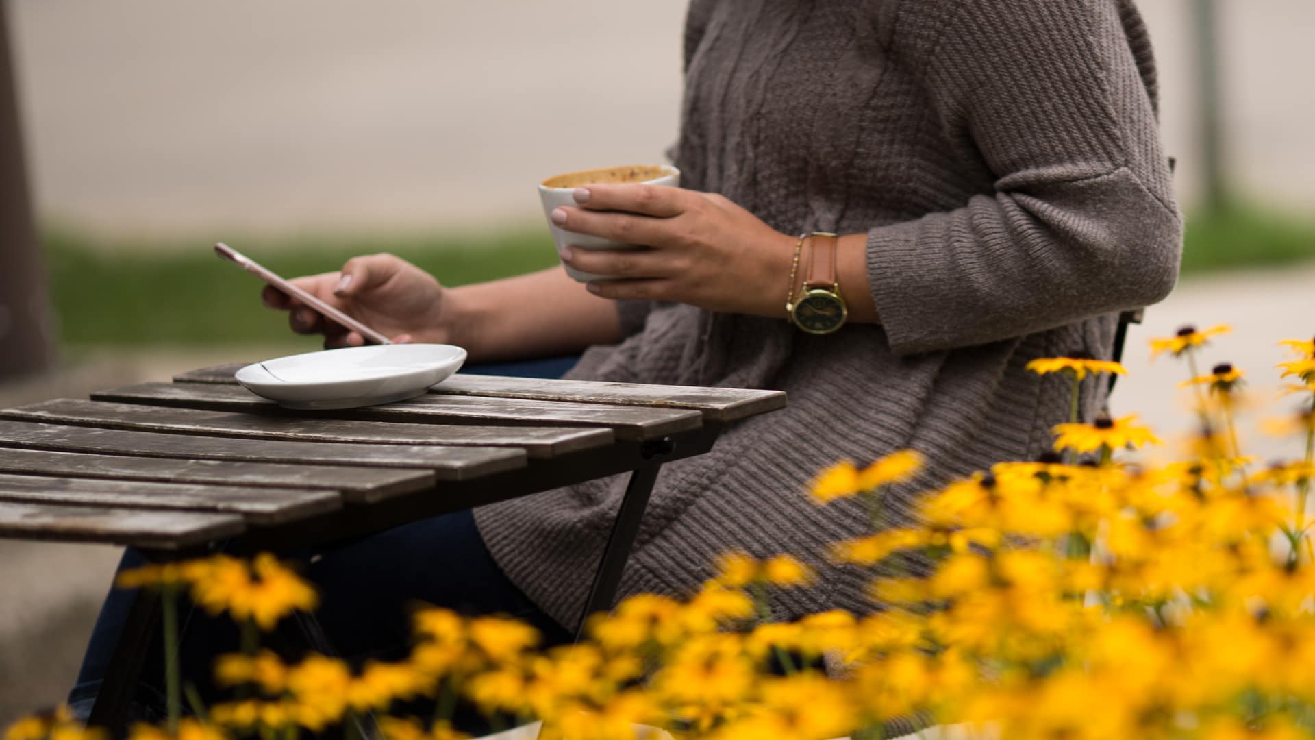 Make-A-Deposit A Grace Student sitting outside at a table, drinking coffee and checking their phone.